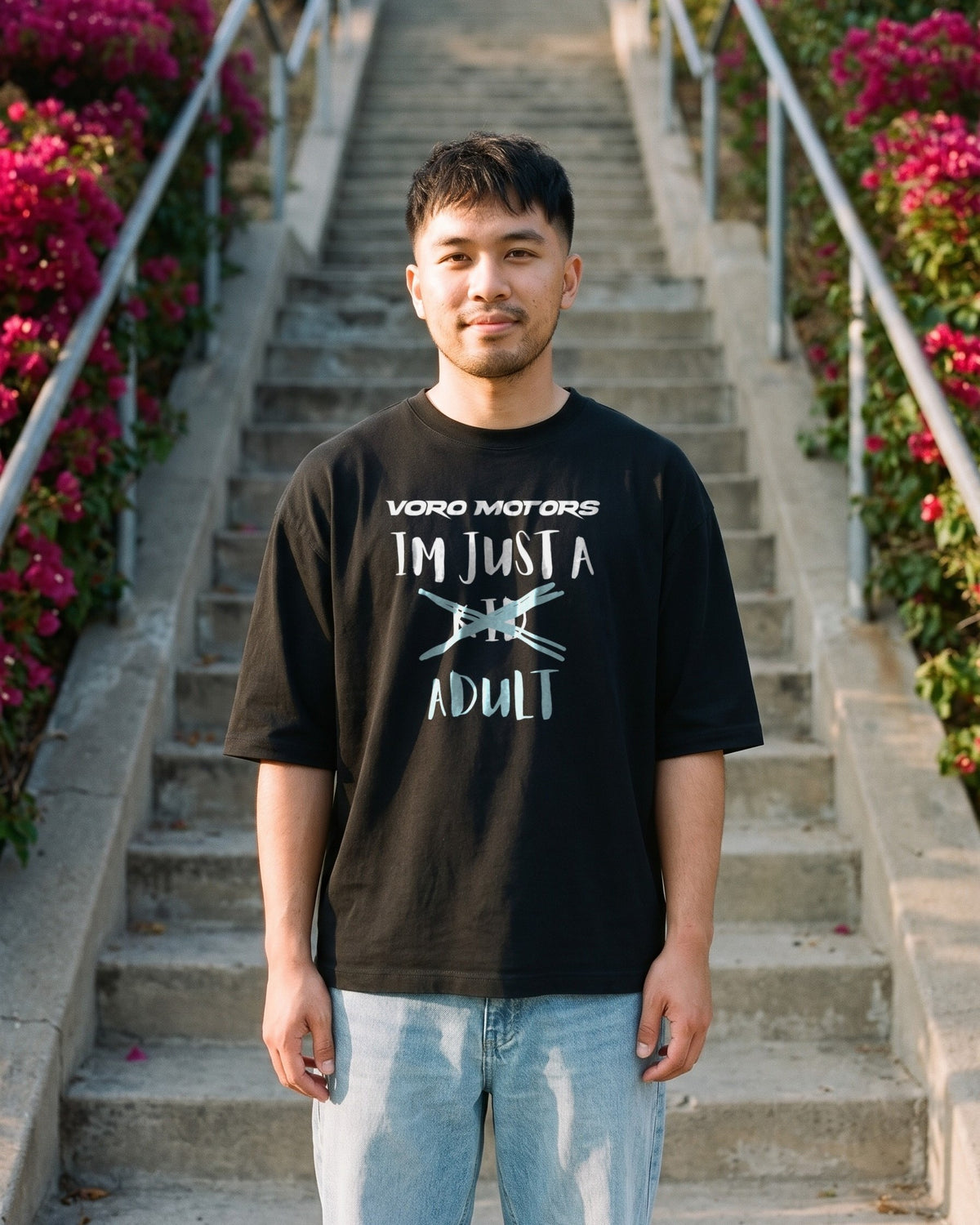Man in the I'm Just a Kid Adult oversized tee on an outdoor stairway with bougainvillea