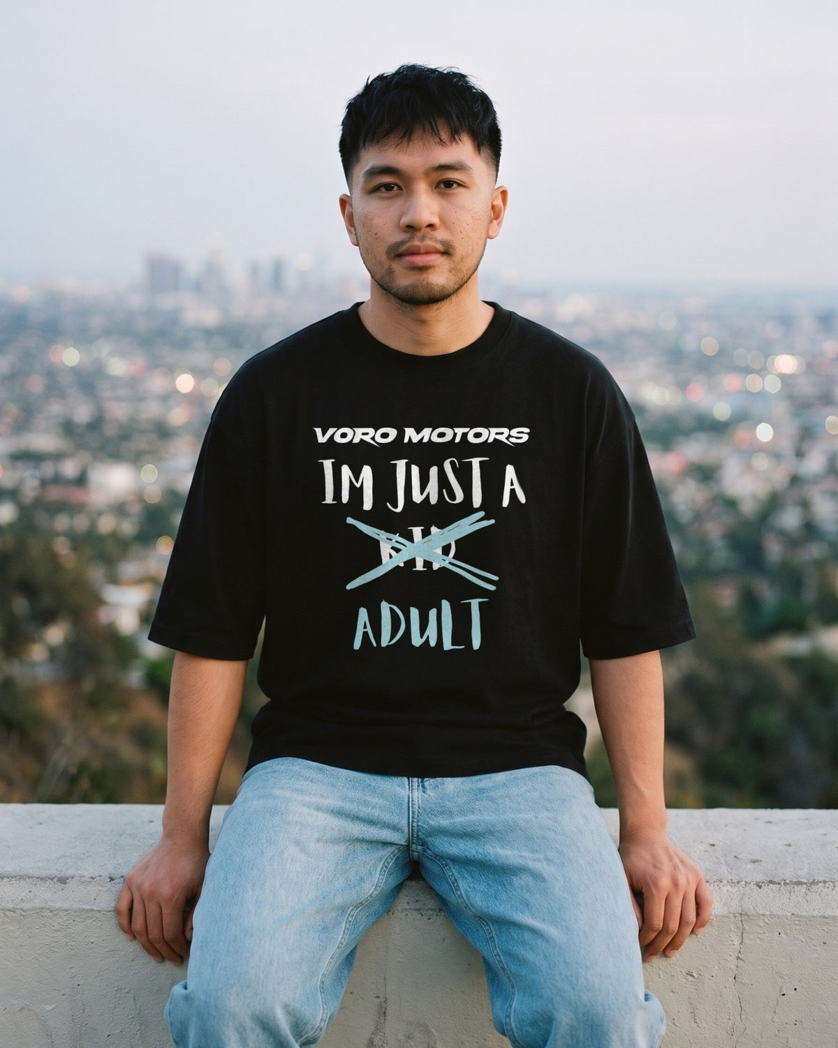 Man in the I'm Just a Kid Adult oversized tee sitting on a ledge above the LA skyline