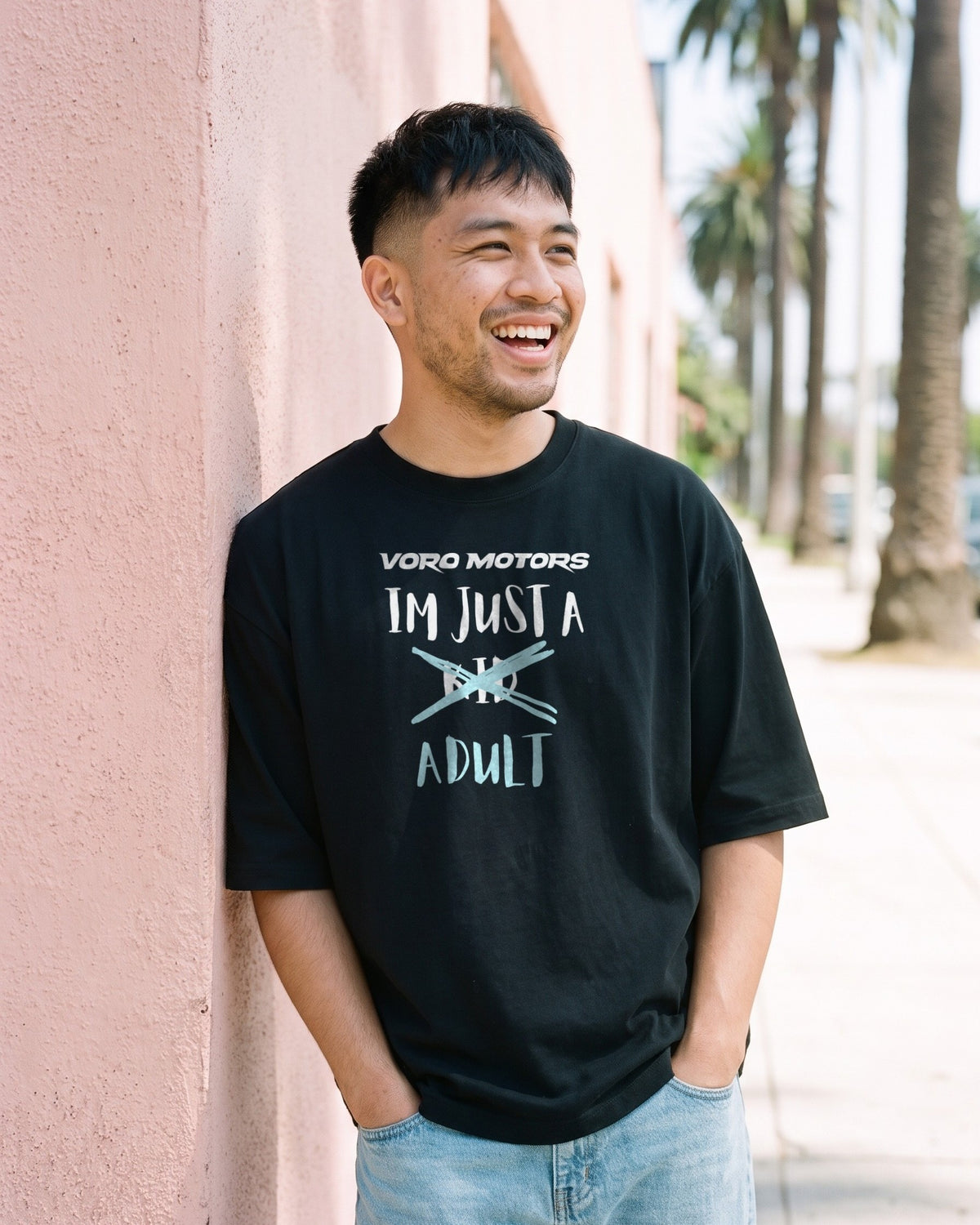 Man laughing in the I'm Just a Kid Adult oversized tee against a pink wall with palms