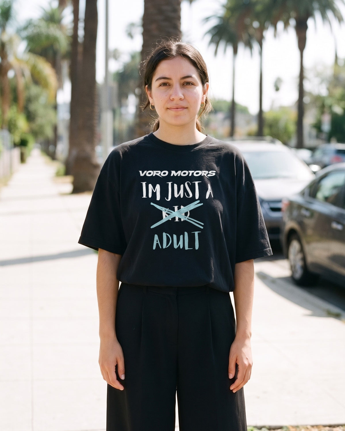Woman wearing the I'm Just a Kid Adult oversized tee in black on a palm-lined street