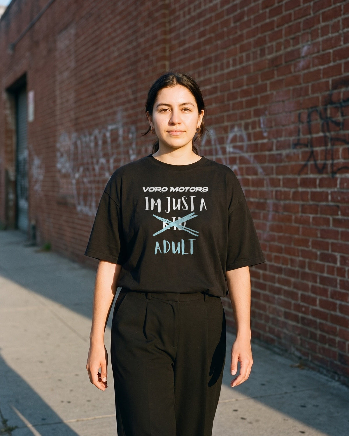 Woman in the I'm Just a Kid Adult oversized tee against a graffiti brick wall