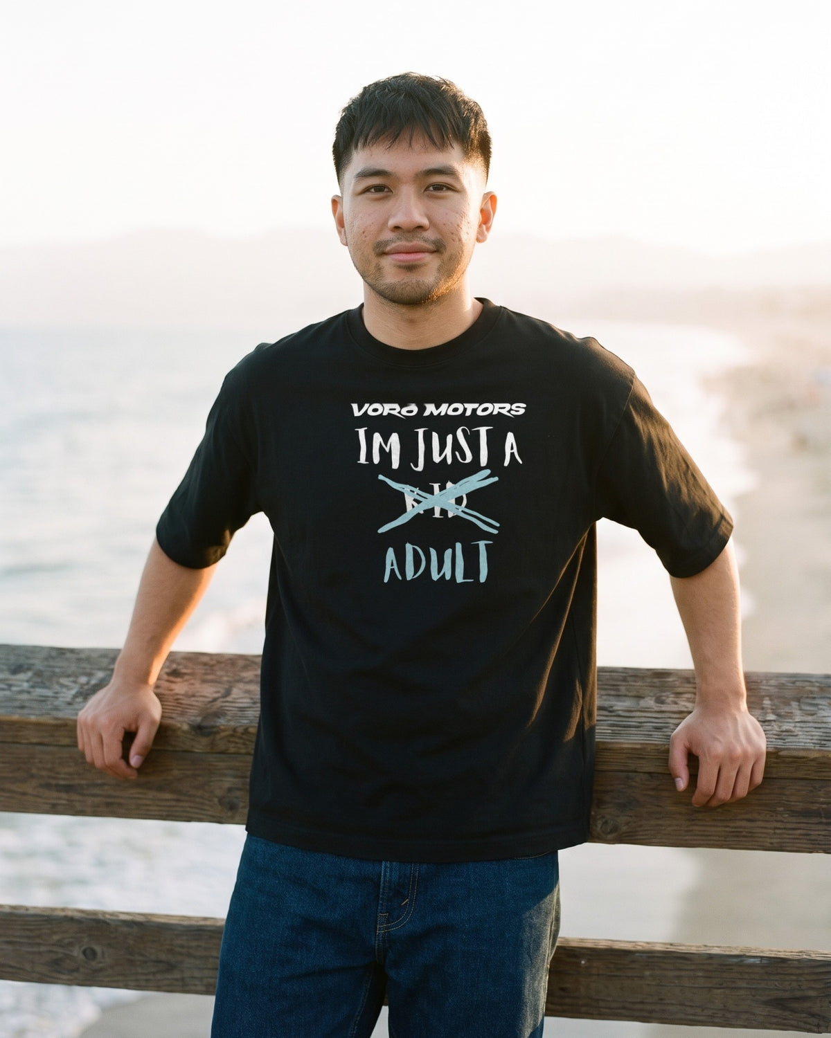 Man in the I'm Just a Kid Adult tee leaning on the Santa Monica Pier rail at sunset